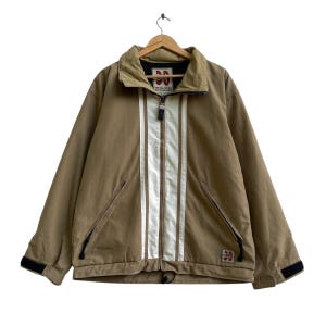 Peut inclure: Veste de couleur beige avec un col beige et une bande blanche sur le devant. La veste a une fermeture éclair, deux poches avant et des détails noirs sur les poignets. Suspendue à un cintre en bois.