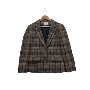 Può includere: Un blazer vintage a quadri nei toni del marrone, grigio e beige, con chiusura a un bottone e due tasche anteriori. La giacca è appesa a una gruccia di legno su sfondo bianco. Il blazer ha un design classico e su misura.