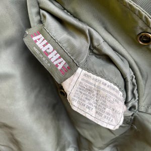 Puede incluir: Primer plano de la etiqueta de una chaqueta Alpha Industries verde oliva. La etiqueta muestra "ALPHA" en rosa y "MADE IN U.S.A.". La etiqueta de la chaqueta proporciona detalles sobre sus especificaciones.