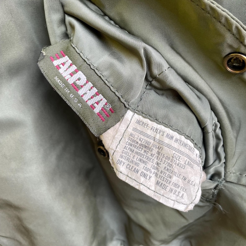 Puede incluir: Primer plano de la etiqueta de una chaqueta Alpha Industries verde oliva. La etiqueta muestra "ALPHA" en rosa y "MADE IN U.S.A.". La etiqueta de la chaqueta proporciona detalles sobre sus especificaciones.