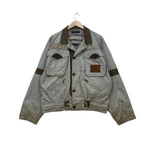 Może przedstawiać: Jasnoszara kurtka z brązowymi akcentami, w tym kieszeniami i kołnierzem. Kurtka ma zapięcie na guziki i talię ze sznurkiem. Brązowe paski na rękawach. Kurtka wisi na drewnianym wieszaku.