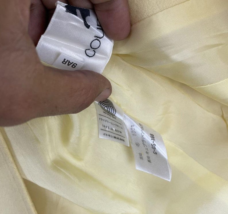 May include: Close-up of a light yellow garment's interior, revealing a tag with the text "COU" and "9AR". Additional tags display Japanese text and the numbers "100%" and "C-DS-168". The garment's lining is a similar pale yellow.