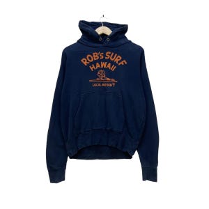 May include: Navy blue hoodie with a kangaroo pocket and drawstring hood. The front features the text "Rob's Surf Hawaii" in orange, with a surfer graphic and the words "Local Motion" below. A casual, vintage-style sweatshirt.