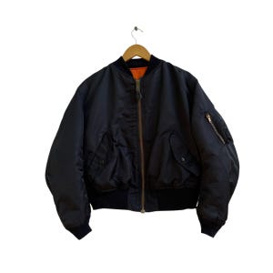Puede incluir: Chaqueta bomber negra con cremallera marrón y forro naranja. La chaqueta tiene dos bolsillos delanteros, un bolsillo con cremallera en la manga izquierda y puños y dobladillo negros de canalé. Cuelga de una percha de madera.