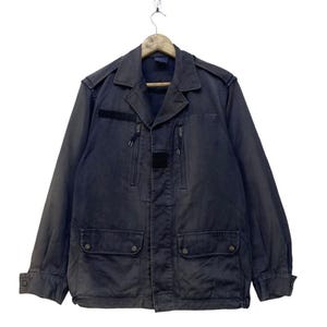 Puede incluir: Chaqueta azul marino oscuro con cuello, dos bolsillos en el pecho y dos bolsillos inferiores. La chaqueta tiene cierre de cremallera y un pequeño parche en la parte superior izquierda del pecho. La chaqueta está colgada de una percha de madera.