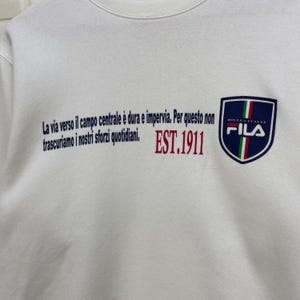 Puede incluir: Sudadera blanca con texto azul y rojo. El texto dice: "La via verso il campo centrale &egrave; dura e impervia. Per questo non trascuriamo i nostri sforzi quotidiani. EST.1911". Un escudo azul marino con la palabra "FILA" est&aacute; a la derecha.