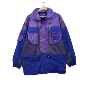 Peut inclure: Veste vintage violette et bleue avec plusieurs poches et un col montant. La veste présente un motif texturé sur la partie supérieure et des panneaux bleus unis sur les manches et la partie inférieure. Elle a une fermeture boutonnée et un cordon de serrage en bas.