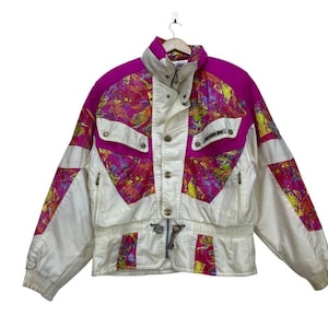 Peut inclure: Veste de ski vintage blanche, fuchsia et panneaux à motifs jaunes, roses et bleus. La veste a un col montant, plusieurs poches et une taille cintrée. La veste est suspendue à un cintre en bois.