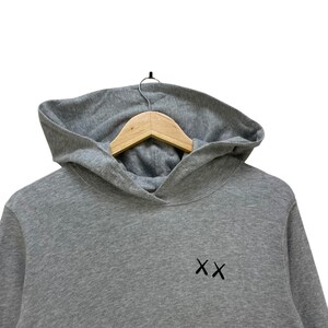 Peut inclure: Un sweat &agrave; capuche gris avec une capuche et des manches longues. Le sweat-shirt porte les lettres brod&eacute;es "XX" sur le devant. Il est suspendu &agrave; un cintre en bois.