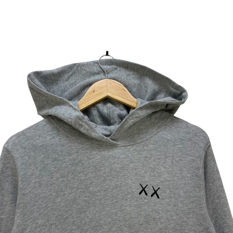 Peut inclure: Un sweat &agrave; capuche gris avec une capuche et des manches longues. Le sweat-shirt porte les lettres brod&eacute;es "XX" sur le devant. Il est suspendu &agrave; un cintre en bois.