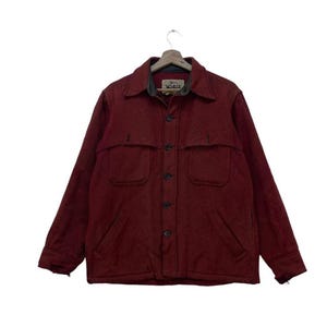 Puede incluir: Chaqueta abotonada burdeos con dos bolsillos en el pecho y dos bolsillos inferiores. La chaqueta tiene cuello y está colgada de una percha de madera. La marca "Woolrich" es visible en el cuello.