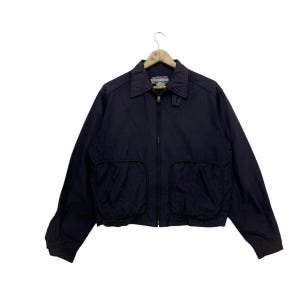 Może przedstawiać: Granatowa kurtka zapinana na zamek błyskawiczny z dwoma dużymi kieszeniami z przodu. Kurtka ma metkę "Wrangler" po lewej stronie kołnierza.