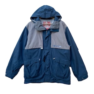May include: A hooded, two-tone jacket in navy blue and gray. The jacket features a full zipper, multiple pockets, and a logo patch on the chest. The hood is attached and the jacket appears to be made of a durable, weather-resistant material.