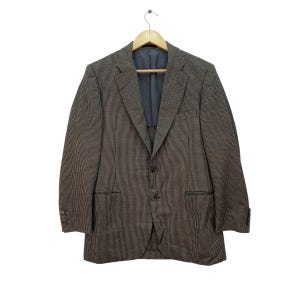 May include: A brown and white houndstooth blazer with a two-button closure, two front pockets, and buttoned cuffs. The blazer is hanging on a wooden hanger against a white background. The interior lining is a dark blue color.