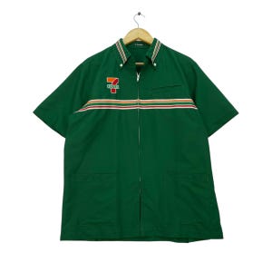 Puede incluir: Camisa verde de manga corta con cremallera completa y dos bolsillos delanteros. La camisa presenta el logotipo "7-Eleven" en el pecho y rayas horizontales en rojo, naranja y blanco. El cuello tiene un diseño a rayas.