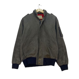 Può includere: Giacca bomber verde oliva con orlo e polsini a coste blu navy. La giacca ha una chiusura a zip, due tasche frontali e una piccola tasca sulla manica. La fodera interna è rossa.