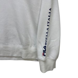 Puede incluir: Sudadera blanca con el logo de Fila en rojo y azul marino. La manga lleva el texto "BIELLA ITALIA" en azul marino. La sudadera tiene cuello redondo y pu&ntilde;os acanalados.