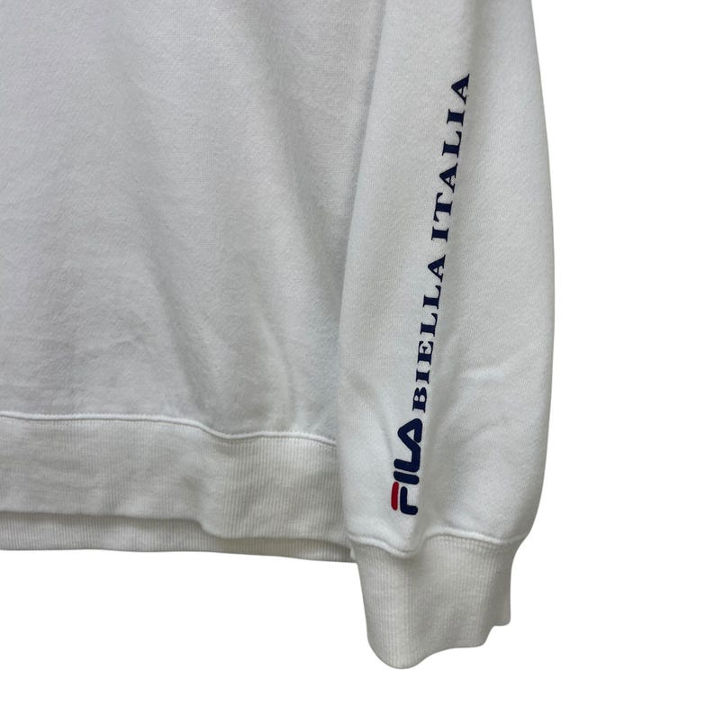 Puede incluir: Sudadera blanca con el logo de Fila en rojo y azul marino. La manga lleva el texto "BIELLA ITALIA" en azul marino. La sudadera tiene cuello redondo y pu&ntilde;os acanalados.