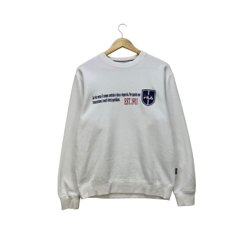 Puede incluir: Sudadera blanca con el texto "La via verso il campo centrale &egrave; dura e imperia. Per questo non trasciniamo i nostri sogni quotidiani." y un logotipo de FILA. La sudadera tiene cuello redondo y mangas largas.