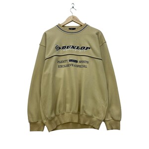 May include: A tan-coloured sweatshirt featuring the "Dunlop Motorsport" logo and the text "Plenty Spirits Exclusive & Special". It has a crew neck with a blue and tan stripe.