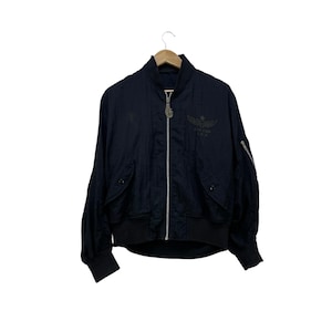 May include: A black bomber jacket with a full-length zip, ribbed cuffs, and a matching hem. The jacket has a small emblem on the chest and a zip pocket on the left sleeve. The jacket is hanging on a wooden hanger.