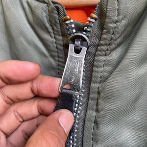 Puede incluir: Primer plano de una cremallera de chaqueta verde oliva con un tirador de metal plateado. El tirador de la cremallera tiene la palabra "IDEAL" estampada. El interior de la chaqueta es naranja.