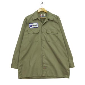 May include: A long-sleeve button-down work shirt in a light olive green color. The shirt features a pointed collar, two chest pockets, and a rectangular patch on the right chest that reads Weather Engineering. A small Dickies brand label is at the hem.