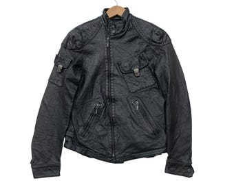 Puede incluir: Chaqueta de cuero negra con cuello alzado, múltiples bolsillos y cremallera. La chaqueta tiene un aspecto texturizado y presenta costuras decorativas en los hombros. La chaqueta está colgada de una percha de madera.