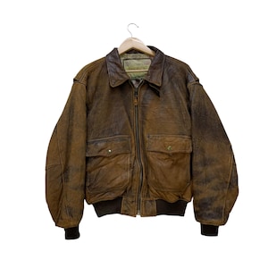 May include: A brown leather bomber jacket with a distressed finish, featuring a zip-up front, two front pockets with button closures, and a ribbed hem and cuffs. The jacket is hanging on a wooden hanger against a white background.