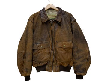 May include: A brown leather bomber jacket with a distressed finish, featuring a zip-up front, two front pockets with button closures, and a ribbed hem and cuffs. The jacket is hanging on a wooden hanger against a white background.