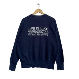 May include: Navy blue sweatshirt with white text on the back. The text reads: "LIFE IS LIKE RIDING A BICYCLE. TO KEEP YOUR BALANCE YOU MUST KEEP MOVING."