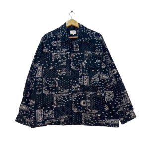 Könnte beinhalten: Ein langärmliges, geknöpftes Hemd mit einem schwarz-weißen Paisley-Muster. Das Hemd hat einen Kragen, zwei Brusttaschen und einen Holzbügel. Der Stoff ist dunkelmarineblau.
