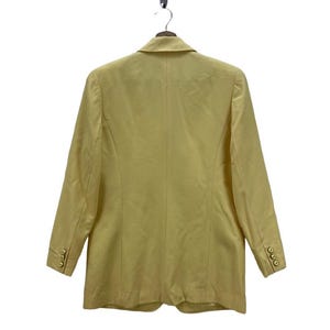 May include: A light yellow blazer with long sleeves, hanging on a hanger. The blazer has a notched lapel, three gold buttons on each cuff, and three gold buttons on the front. The back of the blazer is visible.