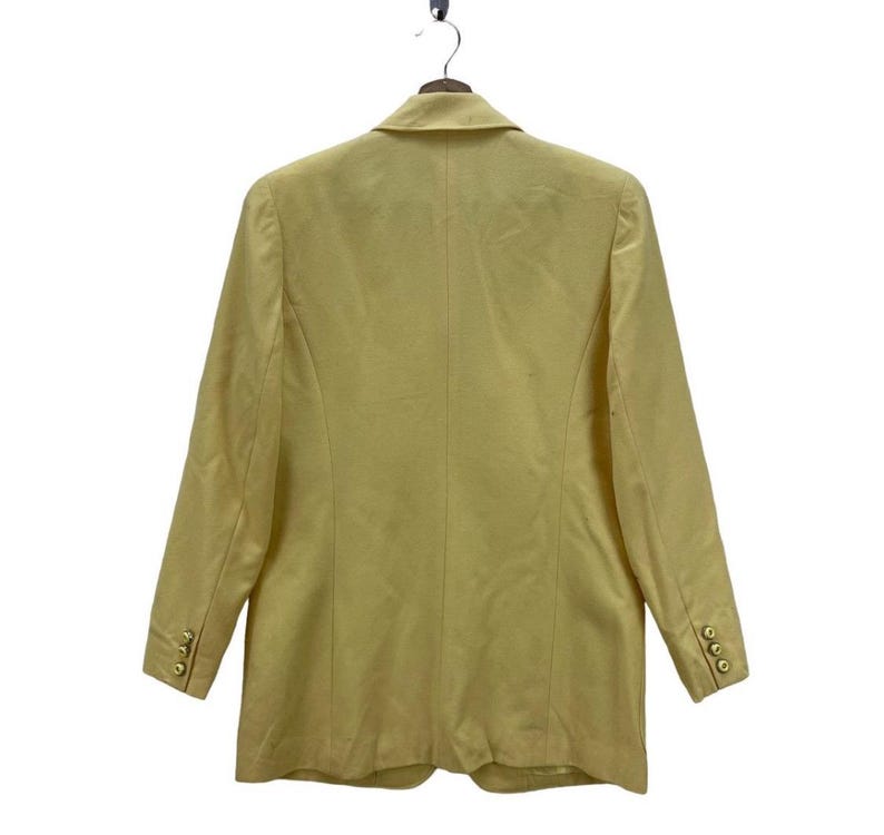 May include: A light yellow blazer with long sleeves, hanging on a hanger. The blazer has a notched lapel, three gold buttons on each cuff, and three gold buttons on the front. The back of the blazer is visible.