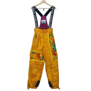 Peut inclure: Salopette de ski jaune avec des motifs floraux et de feuilles. Les bretelles noires ont du texte blanc, une pièce de poitrine bordeaux et une poche zippée. La salopette a des genouillères noires et est suspendue à un cintre en bois.