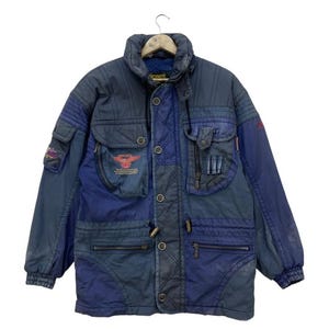 Peut inclure: Veste d'hiver vintage bleue et marine avec plusieurs poches et un col montant. La veste présente une fermeture boutonnée sur le devant, une poche zippée et un écusson avec du texte et un logo. La veste a un design matelassé et des poignets côtelés.