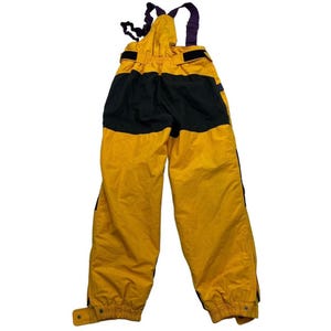 Peut inclure: Salopette de ski jaune et noire avec bretelles réglables. La salopette a une partie supérieure jaune, une partie centrale noire et des jambes jaunes. Les bretelles sont violettes et noires. Fermeture boutonnée à la cheville.