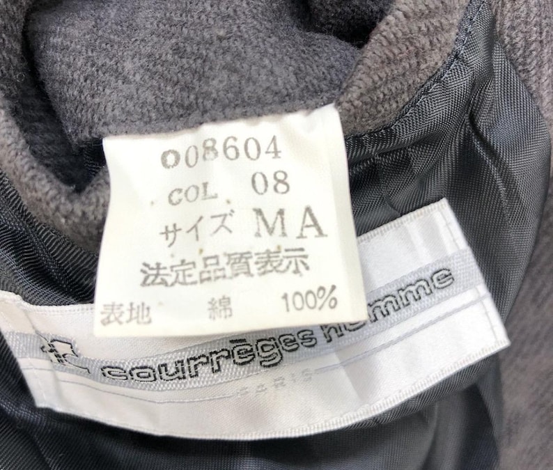 May include: A white tag with black text that reads "008604", "COL. 08", "サイズ MA", "法定品質表示", "綿 100%". Below the tag is a white tag with black text that reads "courreges homme PARIS".