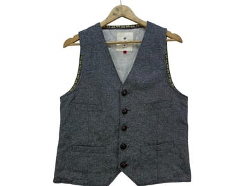 May include: A grey waistcoat with a V-neck and button closure. The waistcoat has two front pockets and a light-coloured lining. It is suspended on a wooden hanger against a white backdrop.