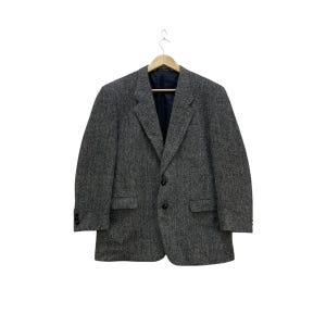 May include: A gray tweed blazer with a herringbone pattern, hanging on a wooden hanger. The blazer has a notched lapel, two front pockets, and a two-button closure. The interior lining is a dark blue color.