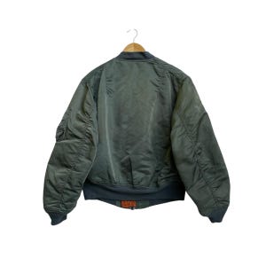 Puede incluir: Chaqueta bomber verde oliva con cuello, pu&ntilde;os y dobladillo de canal&eacute;. La chaqueta tiene un bolsillo con cremallera en la manga izquierda y una peque&ntilde;a etiqueta naranja en la parte inferior. Cuelga de una percha de madera.