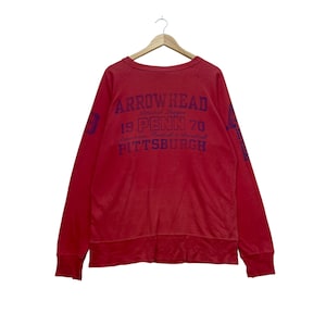 Può includere: Felpa rossa con testo blu sul retro: "ARROWHEAD National League 19 PENN 70 American Football & Baseball PITTSBURGH". La felpa è appesa a una gruccia di legno.
