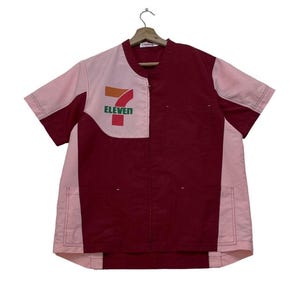 Puede incluir: Una camisa de trabajo de manga corta en color granate y rosa claro, con el logotipo "7-Eleven" en el pecho. La camisa tiene una cremallera frontal y está colgada de una percha de madera. La combinación de colores es una mezcla de granate y rosa claro.