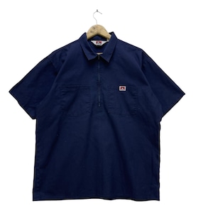 May include: Navy blue short-sleeve shirt with a collar and a quarter-zip closure. The shirt has two front pockets and a small red and white logo patch on the chest. The shirt is hanging on a wooden hanger.