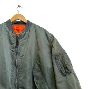 Puede incluir: Chaqueta bomber verde oliva con cuello y pu&ntilde;os grises, colgada de una percha de madera. La chaqueta tiene cierre de cremallera y un bolsillo en la manga izquierda. El forro interior es naranja.