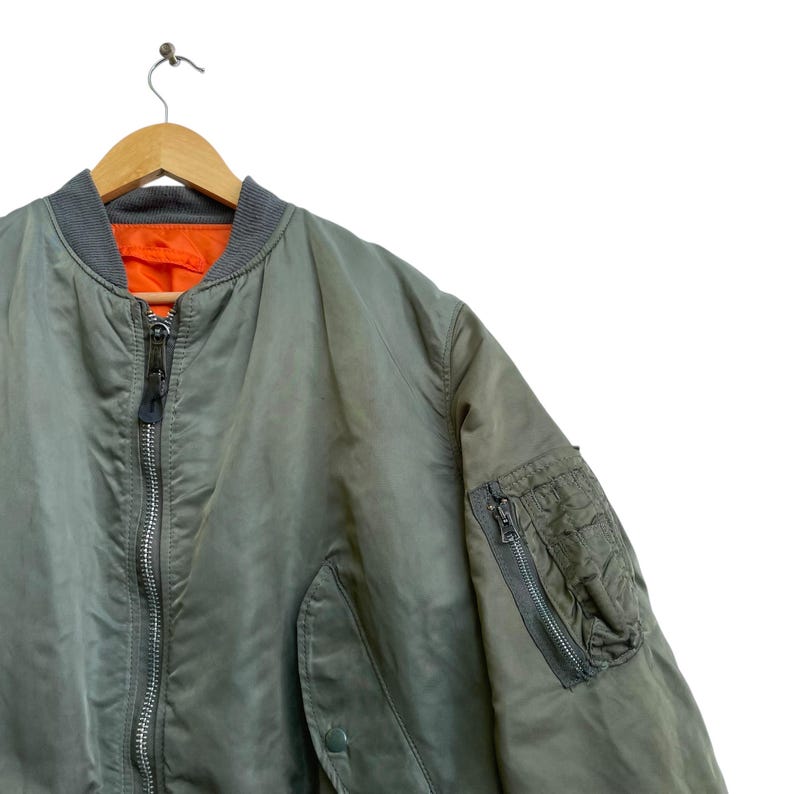 Puede incluir: Chaqueta bomber verde oliva con cuello y pu&ntilde;os grises, colgada de una percha de madera. La chaqueta tiene cierre de cremallera y un bolsillo en la manga izquierda. El forro interior es naranja.