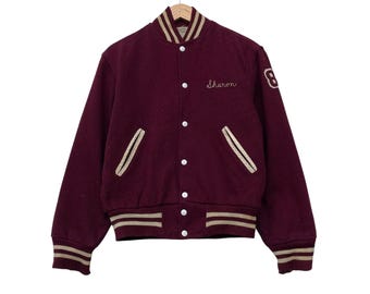 Op de afbeelding: Bordeauxrode varsity jas met beige details. De naam "Sharon" is op de borst geborduurd en het nummer "8" op de mouw. De jas heeft een knoopsluiting, zakken en gestreepte boorden aan de kraag, manchetten en zoom.