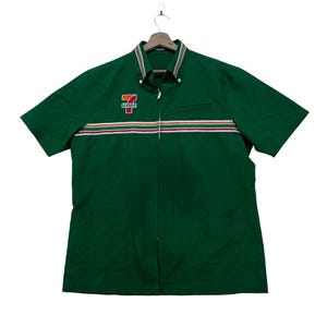 Puede incluir: Camisa de trabajo verde de manga corta con cremallera completa y un pequeño bolsillo. La camisa presenta el logotipo de 7-Eleven en el pecho y rayas horizontales rojas, naranjas y blancas. El cuello tiene un diseño a rayas.