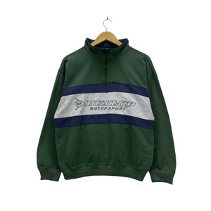Może przedstawiać: Zielona bluza w stylu vintage z granatowo-szarym poziomym paskiem na piersi. Szary pasek zawiera białe logo "DUNLOP MOTOR SPORT". Bluza ma kołnierz z zamkiem błyskawicznym i długie rękawy.