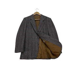 May include: A tweed blazer in shades of brown, gray, and red hangs on a wooden hanger. The jacket features a classic notched lapel, two front pockets, and a brown lining. The blazer is open, revealing the interior.
